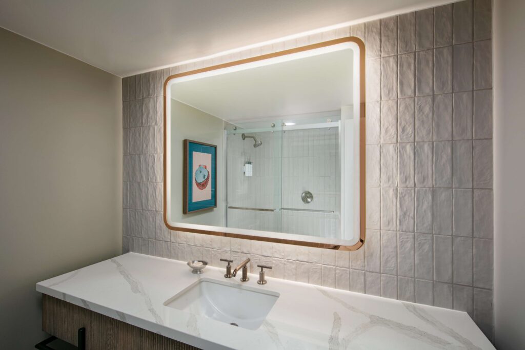 Bathroom vanity with a lit mirror and a shower in the background.